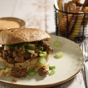 A sloppy joe overflowing from it's bun, with a basket of fries and little bowl of spicy mayo